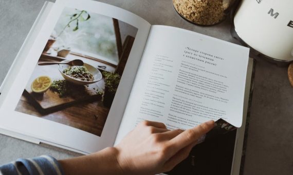 From above of crop faceless woman reviewing article in recipe book in kitchen